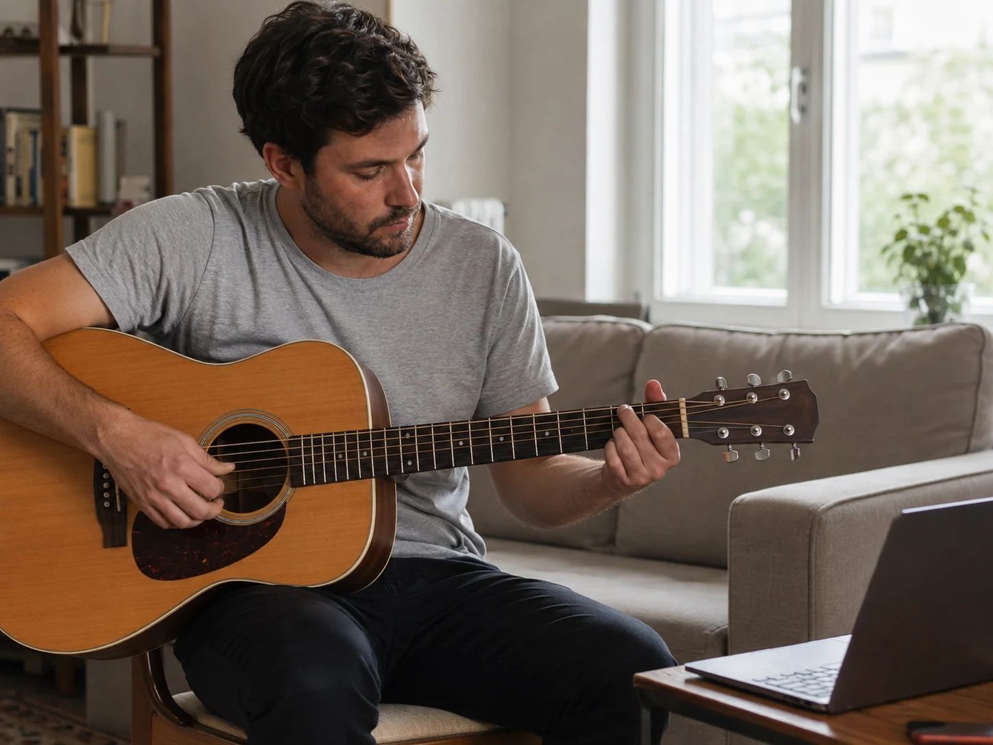 An adult guitarist practices chord voicings beside a laptop in a bright living room.